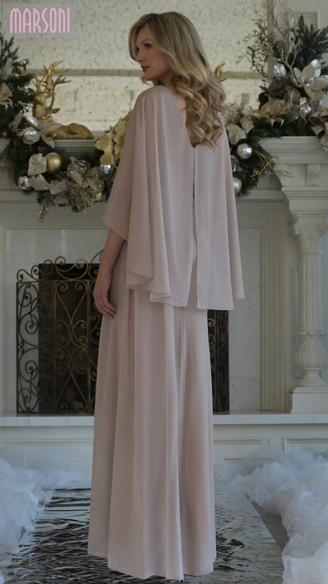 A woman in the Marsoni MV1361 long formal dress, featuring flowing light pink fabric and cape sleeves, stands with her back to the camera before a holiday-decorated fireplace.
